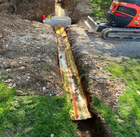 Open trench with ducting and warning tape beside an excavator
