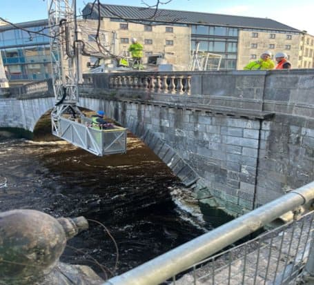 Crew working from a suspended platform on the side of a stone bridge over the River Lee